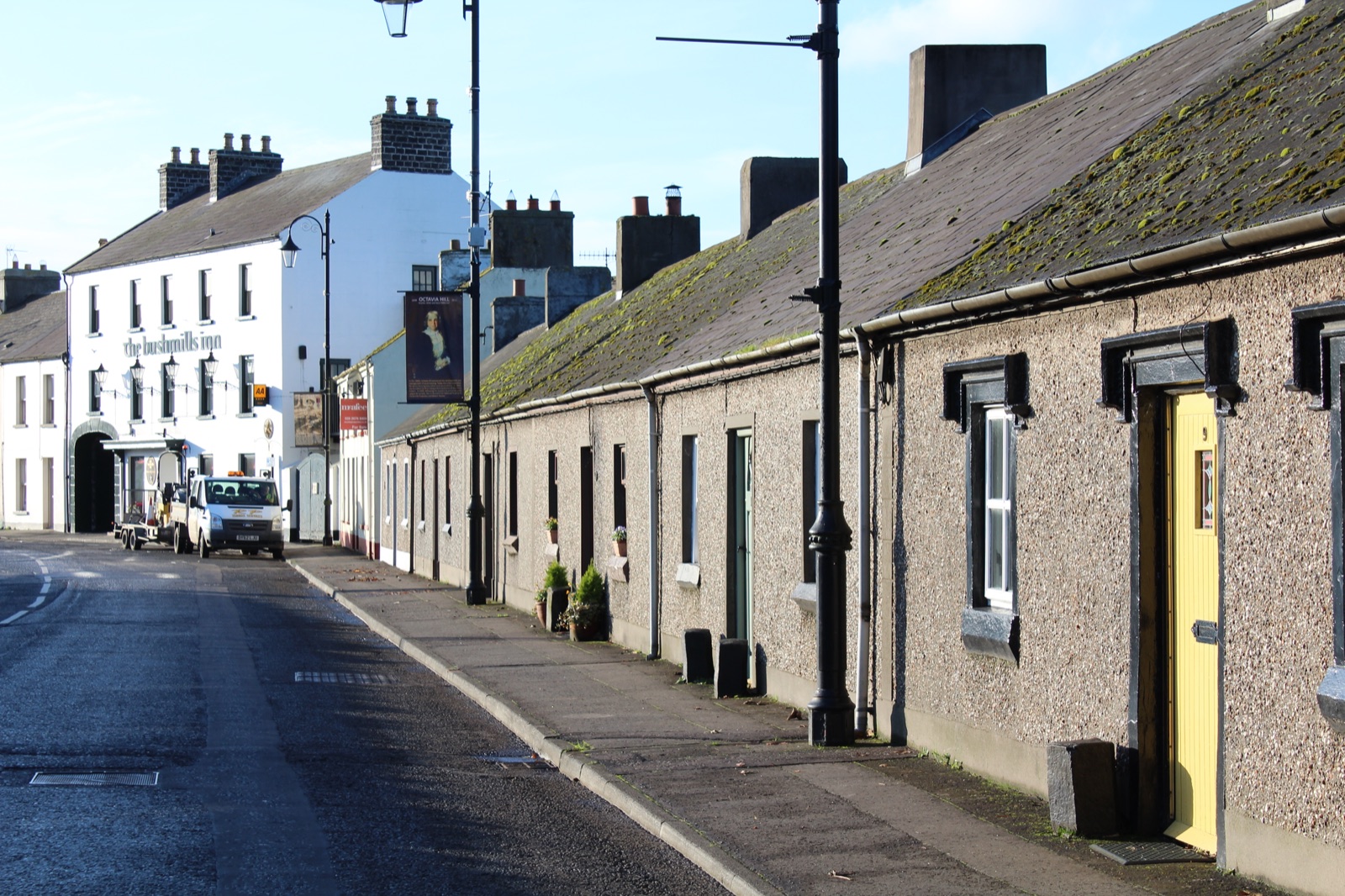 A row of stone cottages on Shell Row in Bushmills, with a yellow front door in the foreground and the Bushmills Inn visible in the distance