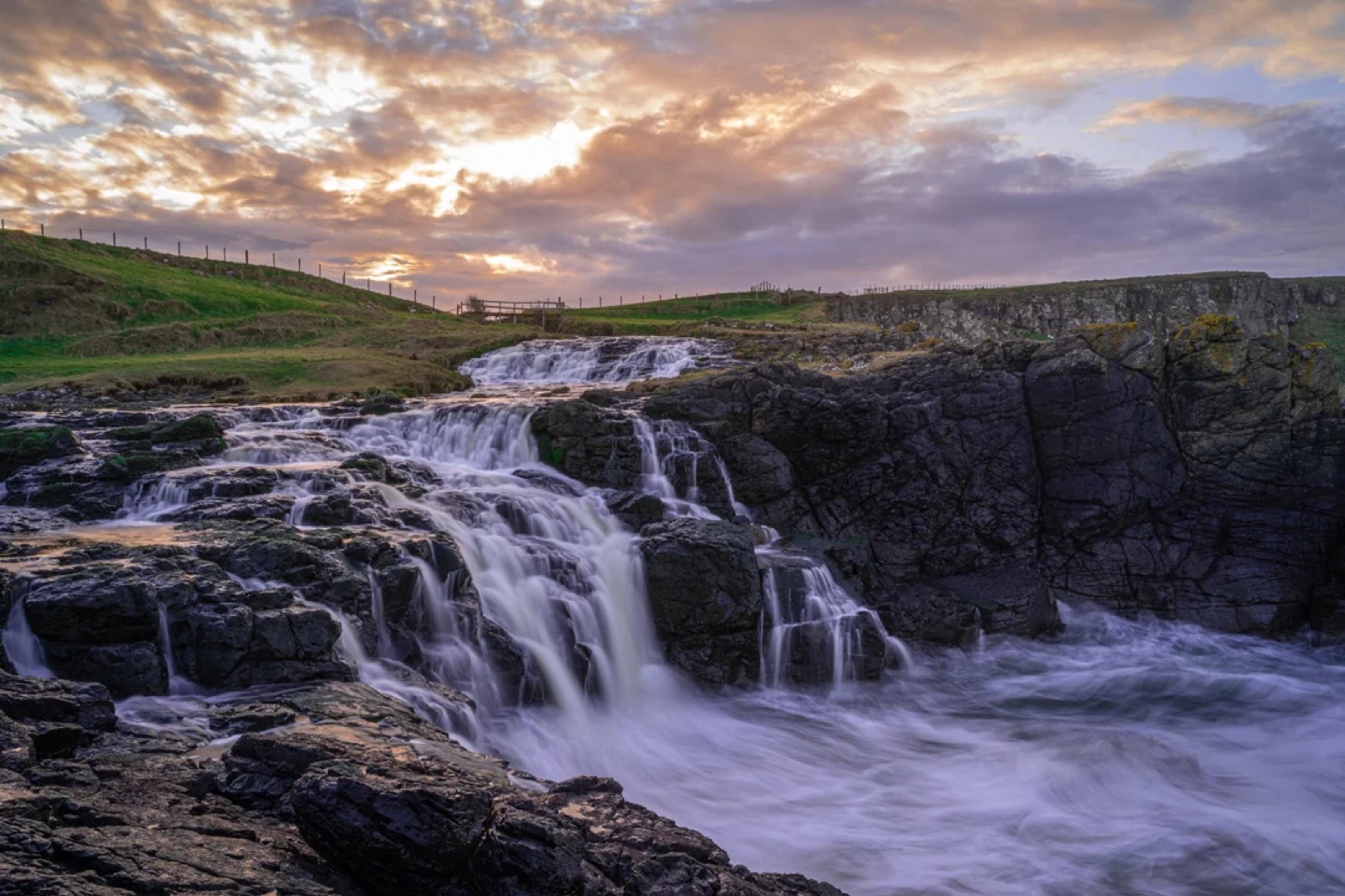 Dunseverick Falls tumbling over dark rock ledges toward the sea at sunset
