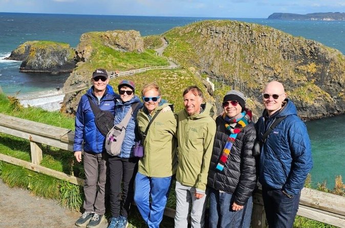 A group of six tourists posing at Carrick-a-Rede with the rope bridge and coastline behind them